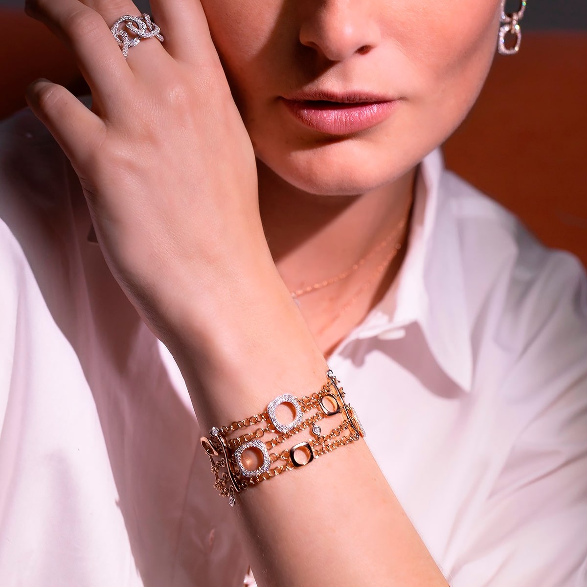 2025 © BIGLI Eine Frau in einem weißen Hemd trägt elegante BIGLI Armschmuck Armbänder aus Gold und Diamanten an ihrem Handgelenk, zusammen mit einem Diamantring und Ohrringen. Ihre Hand ruht sanft auf ihrem Gesicht, was den funkelnden Schmuck noch besser zur Geltung bringt.