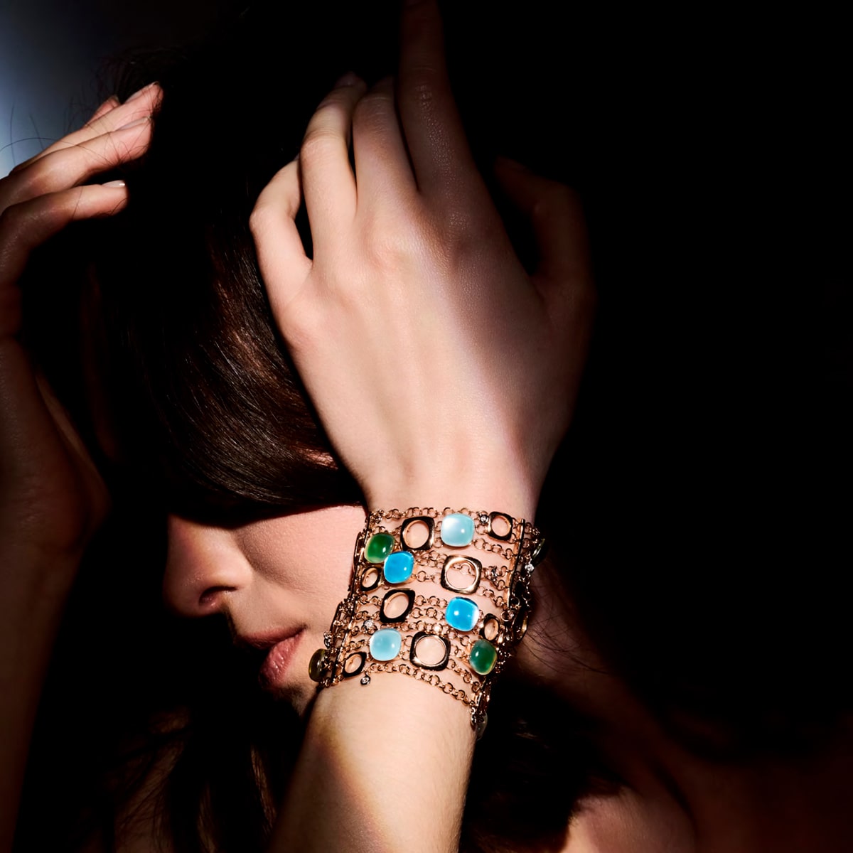 2025 © BIGLI Eine Frau posiert mit der Hand an ihrem Kopf und präsentiert BIGLI Armschmuck: mehrere goldene Armbänder, die mit türkisen, grünen und weißen Steinen verziert sind; dramatische Schatten verdecken teilweise ihr Gesicht.