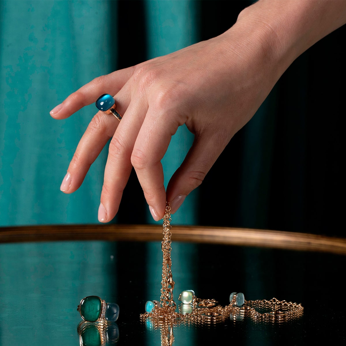 2025 © BIGLI Eine Hand mit einem blauen Edelsteinring hält eine BIGLI Halsschmuck-Goldkette über eine spiegelnde Oberfläche. Weitere Goldketten und Edelsteinringe sind in der Nähe verstreut, im Hintergrund ist ein blaugrüner Vorhang zu sehen.