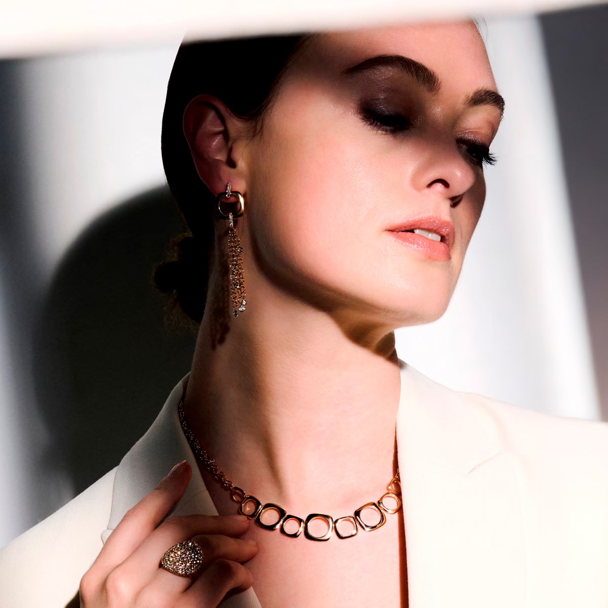 2025 © BIGLI Eine Frau in einem weißen Blazer trägt eine goldene Halskette, BIGLI Ohrringe und einen Ring. Sie blickt mit weichem Make-up nach unten, während dramatisches Licht ihren Schmuck hervorhebt.