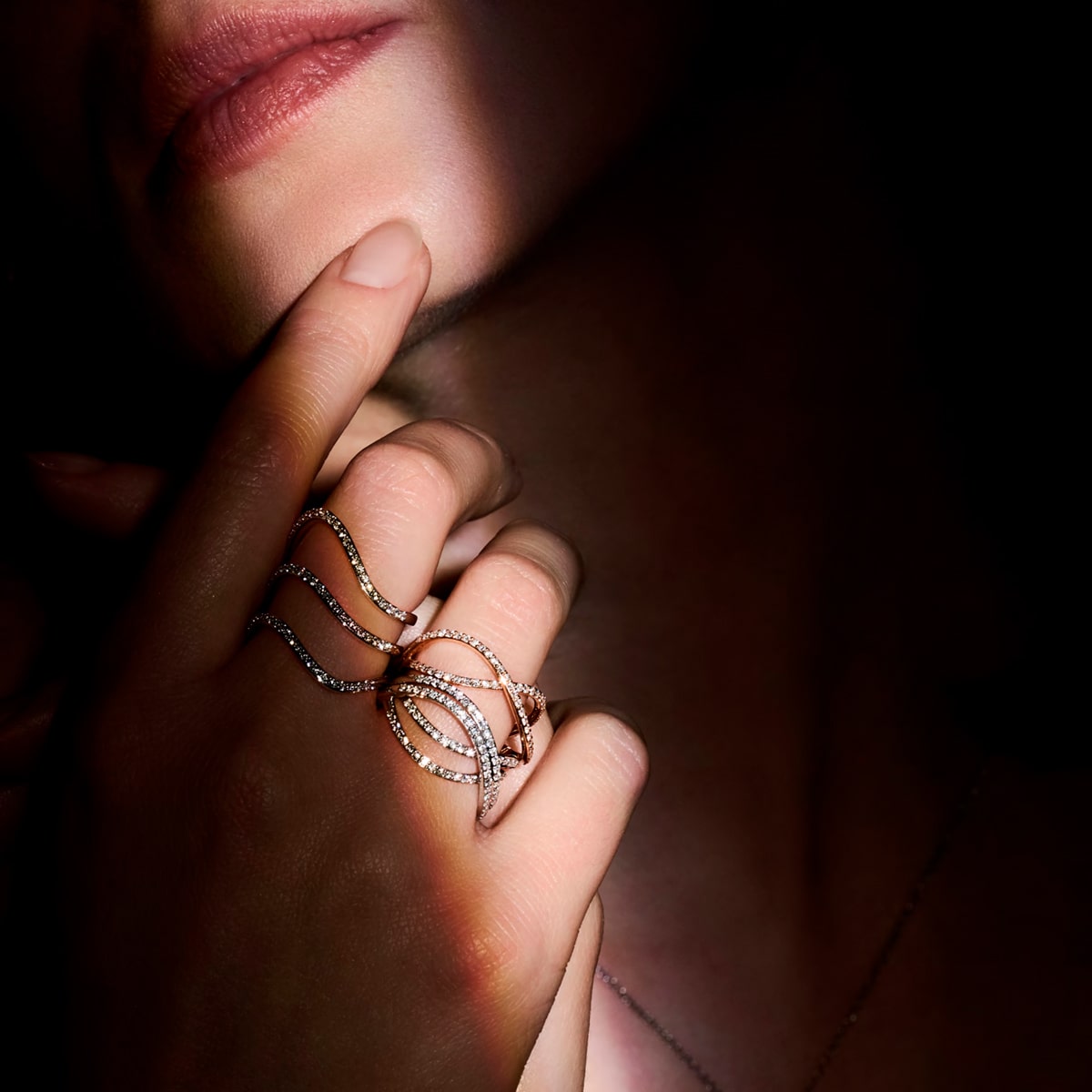 2025 © BIGLI Eine Nahaufnahme der Hand einer Person, die sanft ihr Gesicht berührt, zeigt elegante, gewellte BIGLI Ringe, die mit Diamanten verziert sind. Weiches Licht hebt den Schmuck und die Lippen hervor, während der größte Teil des Bildes im Schatten bleibt.