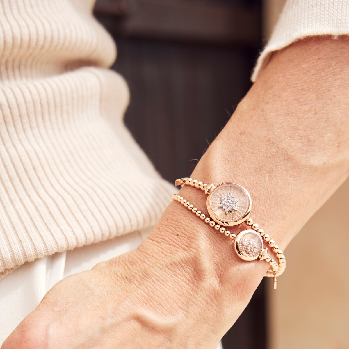 2025 © CAPOLAVORO Nahaufnahme des Handgelenks einer Person, die zwei goldene Armschmuck-Armbänder von Capolavoro trägt, eines davon mit einem Starburst-Muster und einem runden Anhänger, kombiniert mit einem cremefarbenen Rippenpullover.