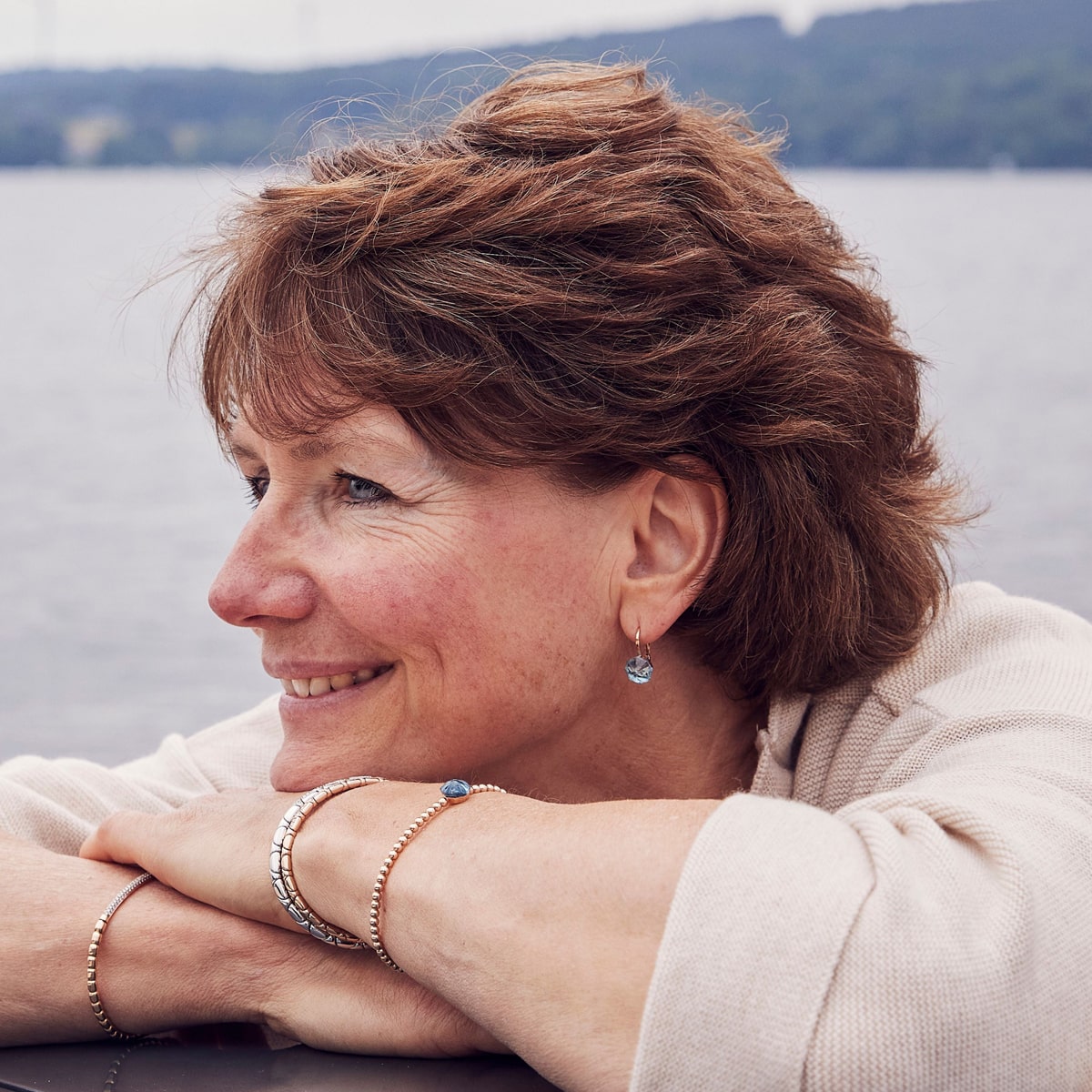 2025 © CAPOLAVORO Eine Frau mit kurzen braunen Haaren lächelt, während sie ihre mit Capolavoro Armschmuck geschmückten Arme auf einer Fläche an einem See ausruht und Ohrringe und Armbänder trägt. Im Hintergrund sind Wasser und ferne, baumbewachsene Hügel zu sehen.