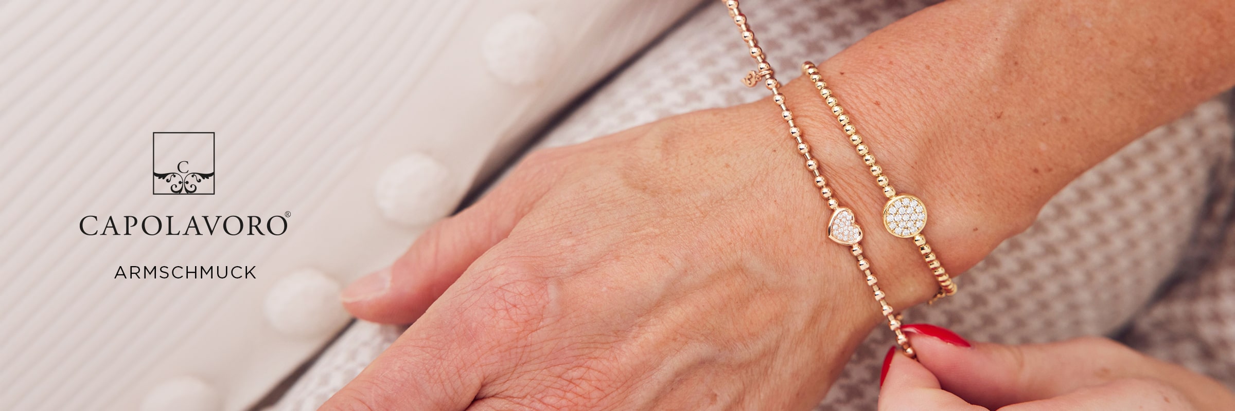 2025 © CAPOLAVORO Nahaufnahme eines Handgelenks, das mit zwei roségoldenen Capolavoro-Armschmuck-Armbändern mit herzförmigen Anhängern geschmückt ist. Die Person verstellt ein Armband vor einem Hintergrund aus strukturiertem Stoff und dem Text "CAPOLAVORO ARMSCHMUCK".