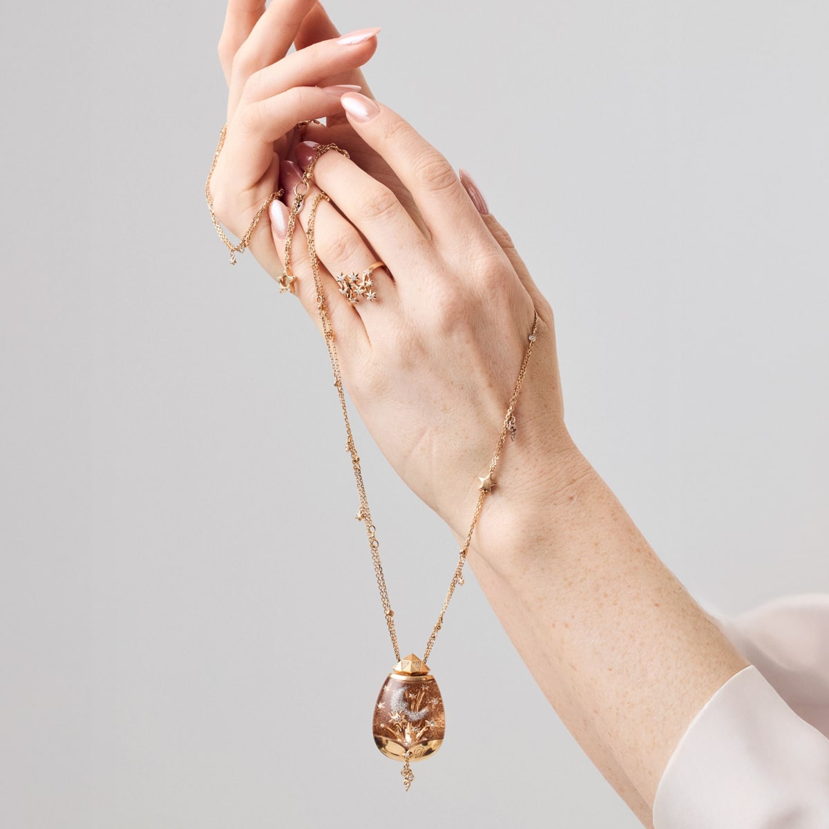 2025 © CAPOLAVORO Zwei Hände halten eine zarte Goldkette von Capolavoro Halsschmuck mit einem großen, verschnörkelten Anhänger. Die Person trägt ein leichtes, langärmeliges Oberteil und einen Goldring mit kleinen Schmucksteinen. Der Hintergrund ist schlicht und sanft beleuchtet.
