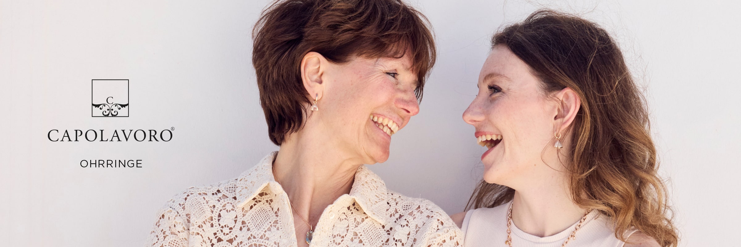 2025 © CAPOLAVORO Zwei Frauen lächeln sich an, beide tragen Spitzenoberteile, eleganten Schmuck und umwerfende Capolavoro Ohrringe. Das CAPOLAVORO-Logo und das Wort "OHRRINGE" erscheinen auf der linken Seite des Bildes.
