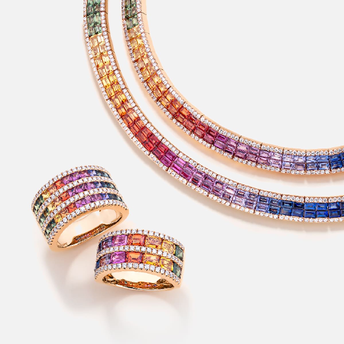 2025 © Diamond Group Zwei goldene Ringe und zwei goldene Halsketten von DiamondGroup sind mit mehrfarbigen Edelsteinen im Quadratschliff besetzt, die in Regenbogenmustern angeordnet sind und auf einem weißen Hintergrund erscheinen.