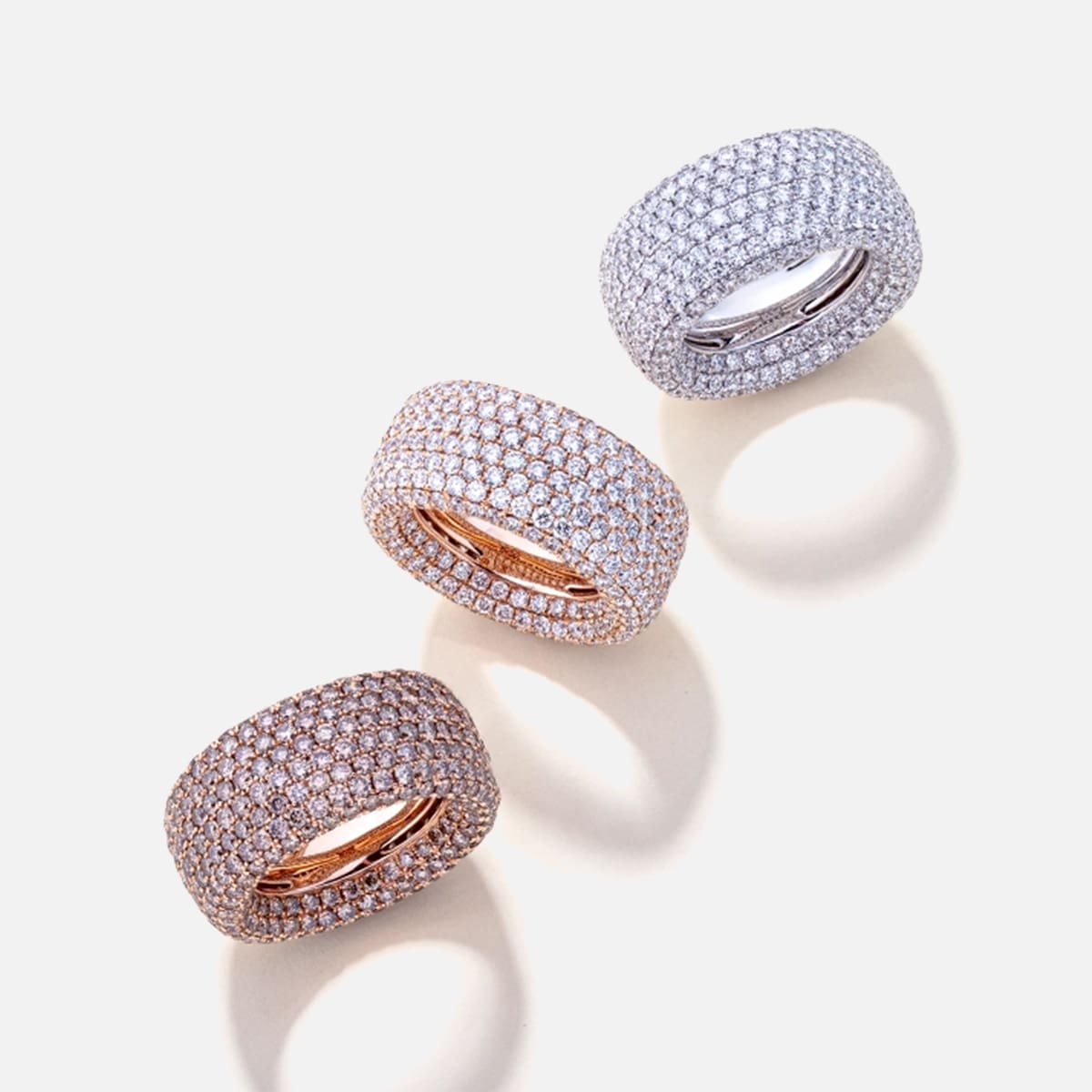 2025 © Diamond Group Drei breite Bandringe der DiamondGroup sind diagonal auf einem hellen Hintergrund angeordnet. Jeder Ring ist mit mehreren Reihen kleiner Diamanten besetzt; zwei sind aus Roségold und einer aus Weißgold und werfen weiche Schatten.