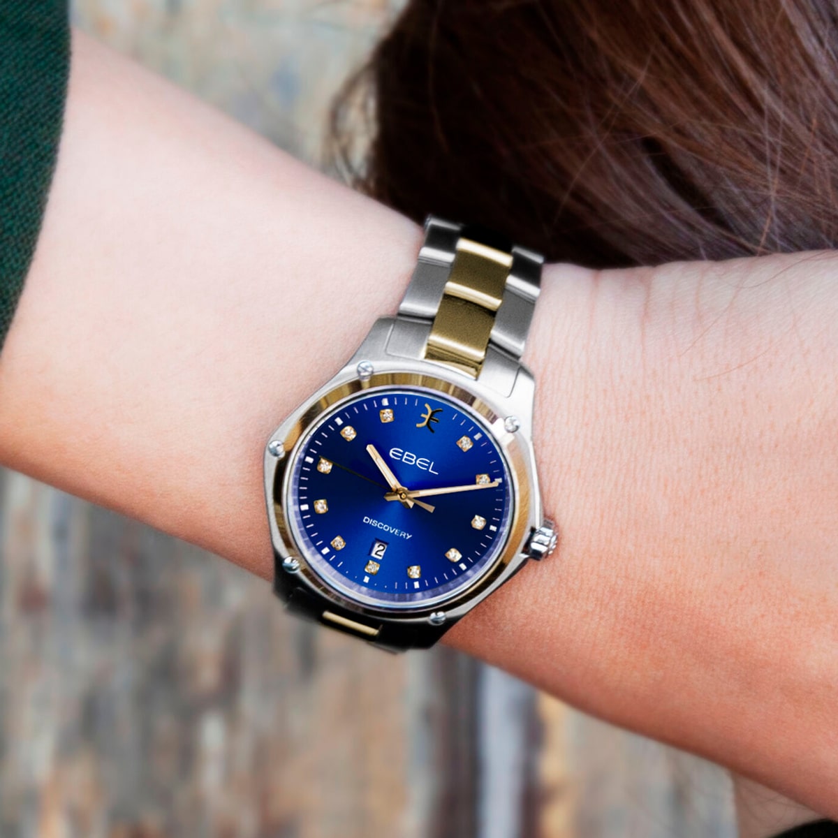 2025 © EBEL Eine Person trägt eine silberne und goldene EBEL Discovery-Armbanduhr mit blauem Zifferblatt und Diamantmarkierungen am Handgelenk vor einem unscharfen Hintergrund.