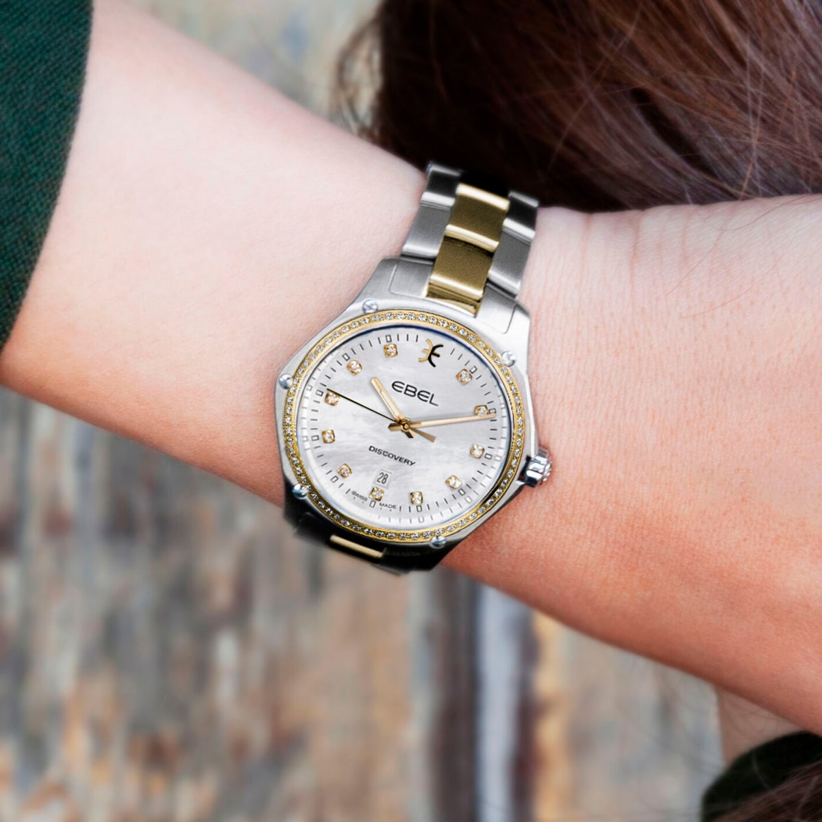 2025 © EBEL Eine Person trägt eine EBEL Discovery Armbanduhr mit einem silbernen und goldenen Metallband und einem runden, weißen Zifferblatt mit goldenen Stundenmarkierungen und Diamantakzenten. Die Uhr befindet sich am linken Handgelenk vor einem unscharfen Hintergrund.