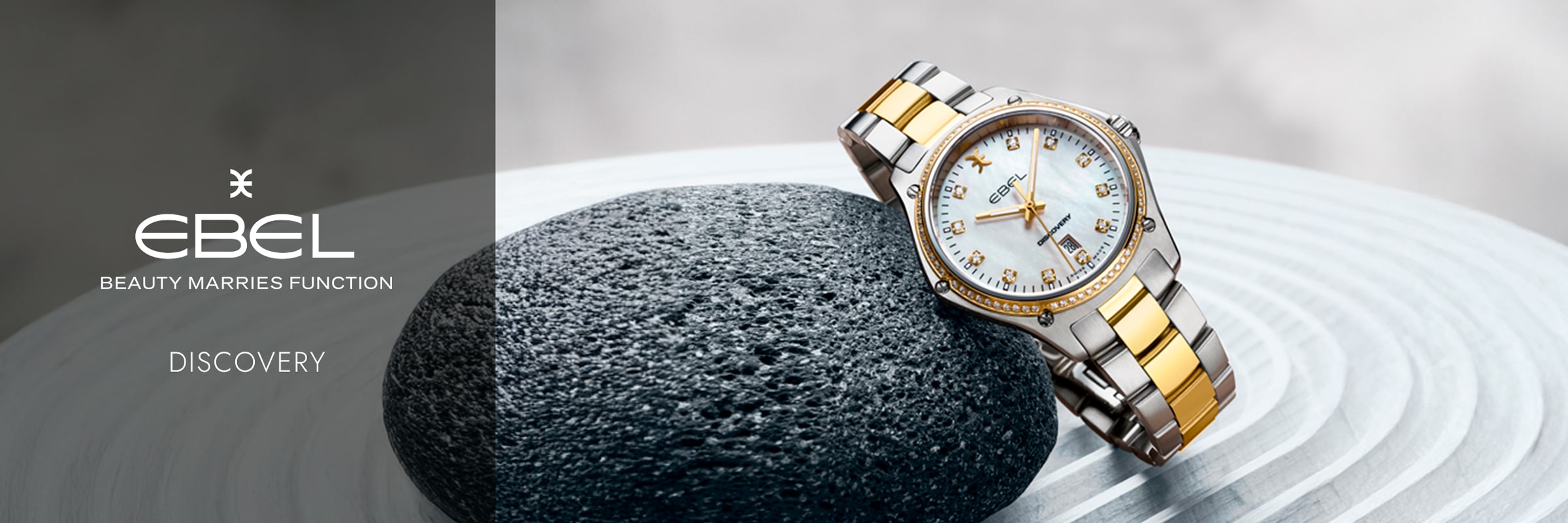 2025 © EBEL Eine luxuriöse EBEL Discovery Uhr mit einem silbernen und goldenen Band ruht auf einem strukturierten schwarzen Stein. Auf dem Hintergrund sind das Logo von Ebel, der Slogan "Beauty Marries Function" und das Wort "Discovery" zu sehen.
