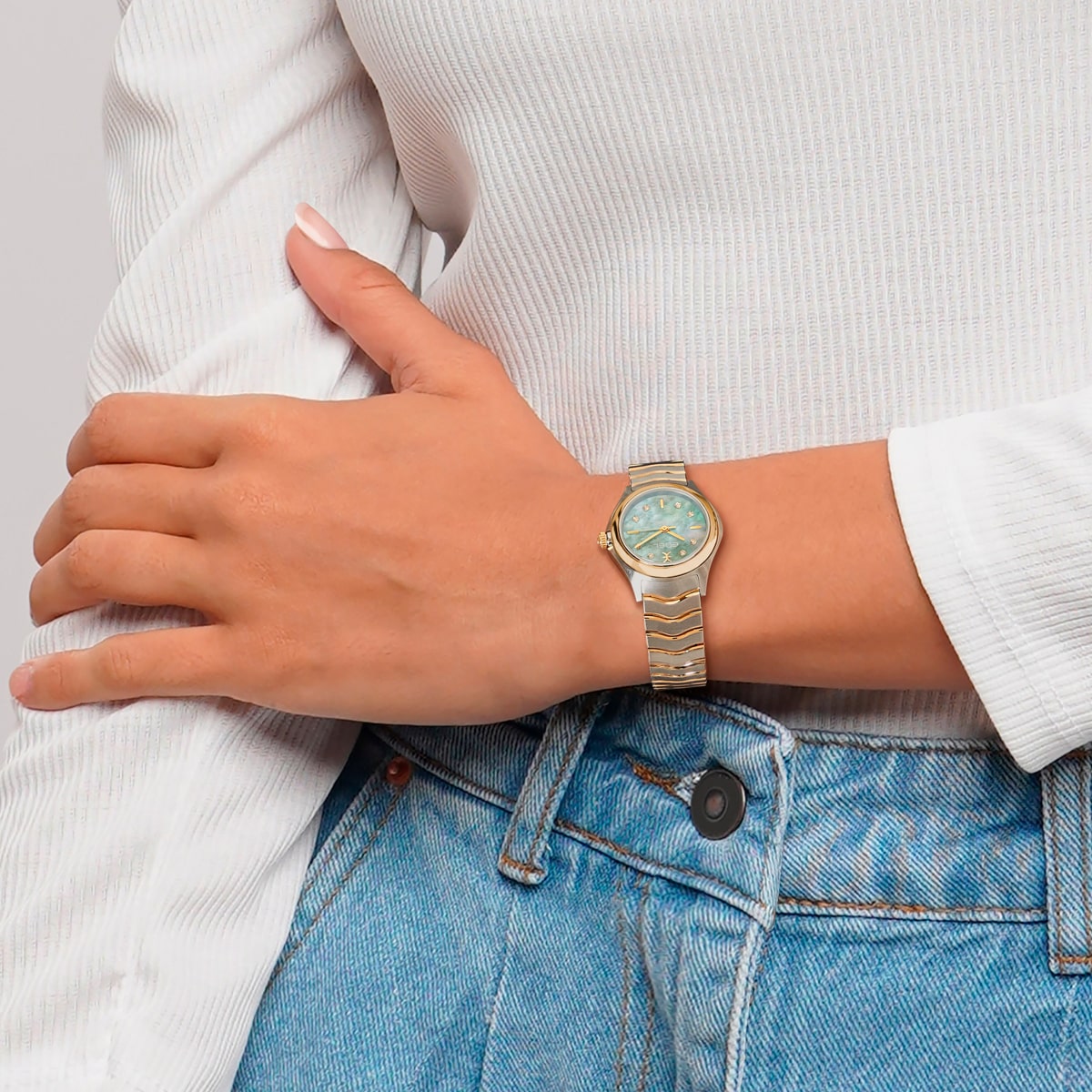 2025 © EBEL Eine Person, die ein weißes langärmeliges Hemd und eine blaue Jeans trägt, hat den linken Arm über dem Körper gekreuzt und zeigt eine goldene EBEL Wave-Armbanduhr mit hellblauem Zifferblatt.
