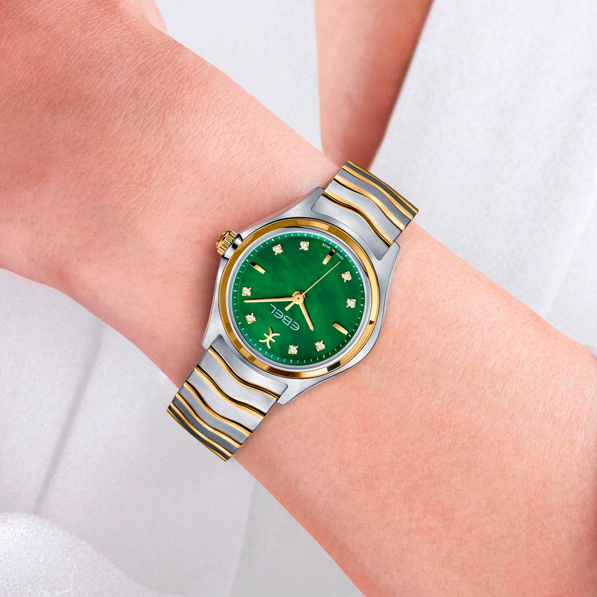 2025 © EBEL Eine Person, die eine silberne und goldene EBEL Wave-Armbanduhr mit grünem Zifferblatt und goldenen Akzenten trägt; die Uhr verfügt über diamantene Stundenmarkierungen und eine goldene Krone, dargestellt vor einem hellen Hintergrund.