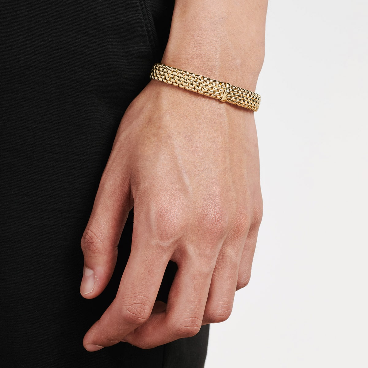 2025 © FOPE Nahaufnahme der Hand und des Handgelenks einer Person, die ein goldenes FOPE-Armschmuck-Armband trägt, wobei die Person in schwarzer Kleidung vor einem schlichten weißen Hintergrund steht.
