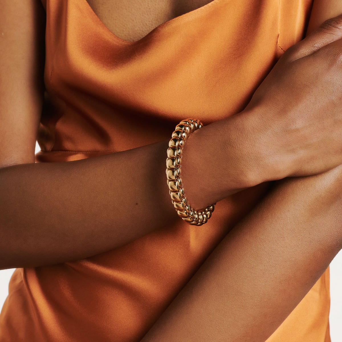 2025 © FOPE Eine Person, die ein ärmelloses, orangefarbenes Oberteil aus Satin trägt, verschränkt die Arme und präsentiert ein klobiges goldenes FOPE-Armschmuck-Armband an ihrem Handgelenk.