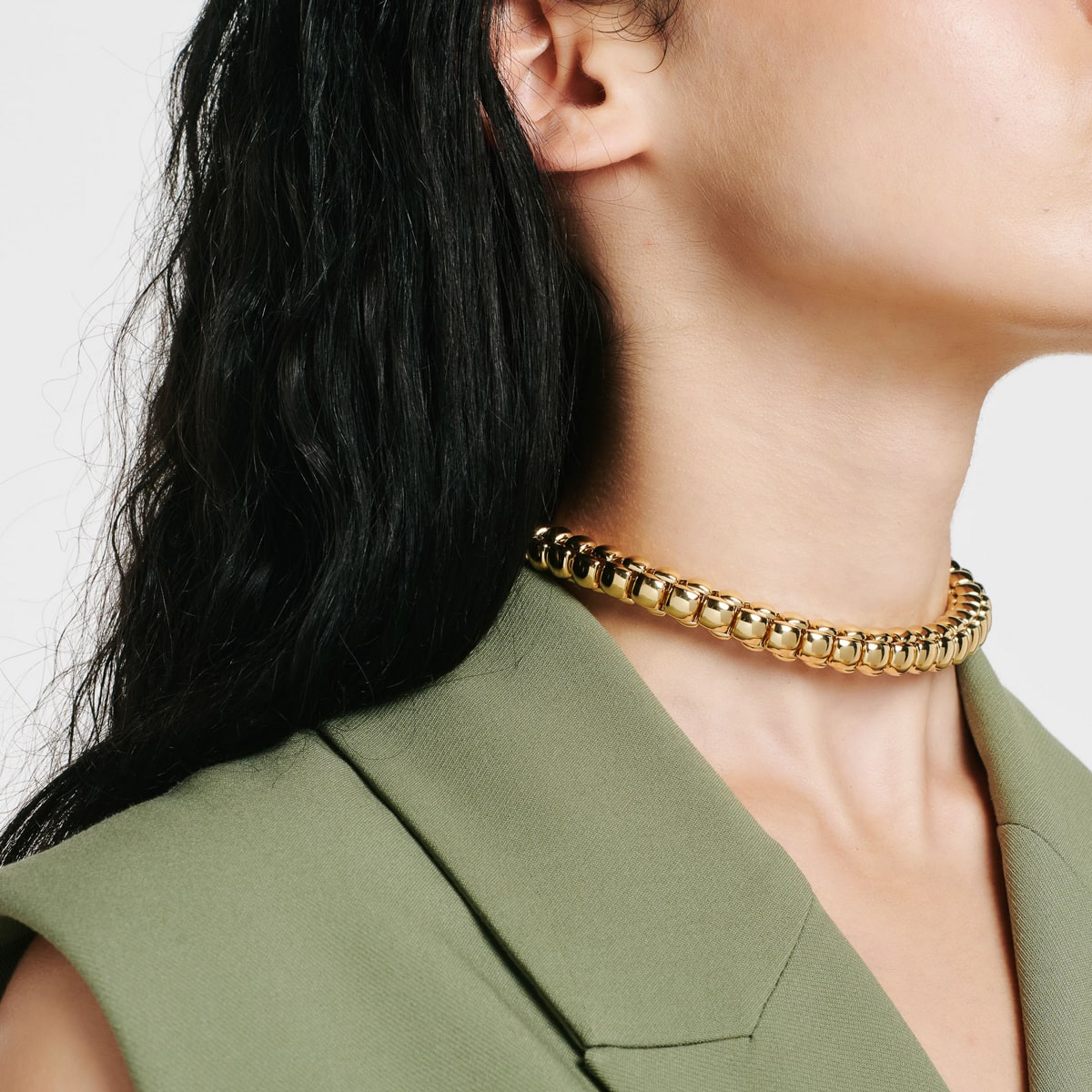 2025 © FOPE Eine Frau mit langen dunklen Haaren, die FOPE Halsschmuck - eine klobige goldene Halskette - und einen hellgrünen Blazer trägt, wird von der Seite vor einem schlichten weißen Hintergrund gezeigt.