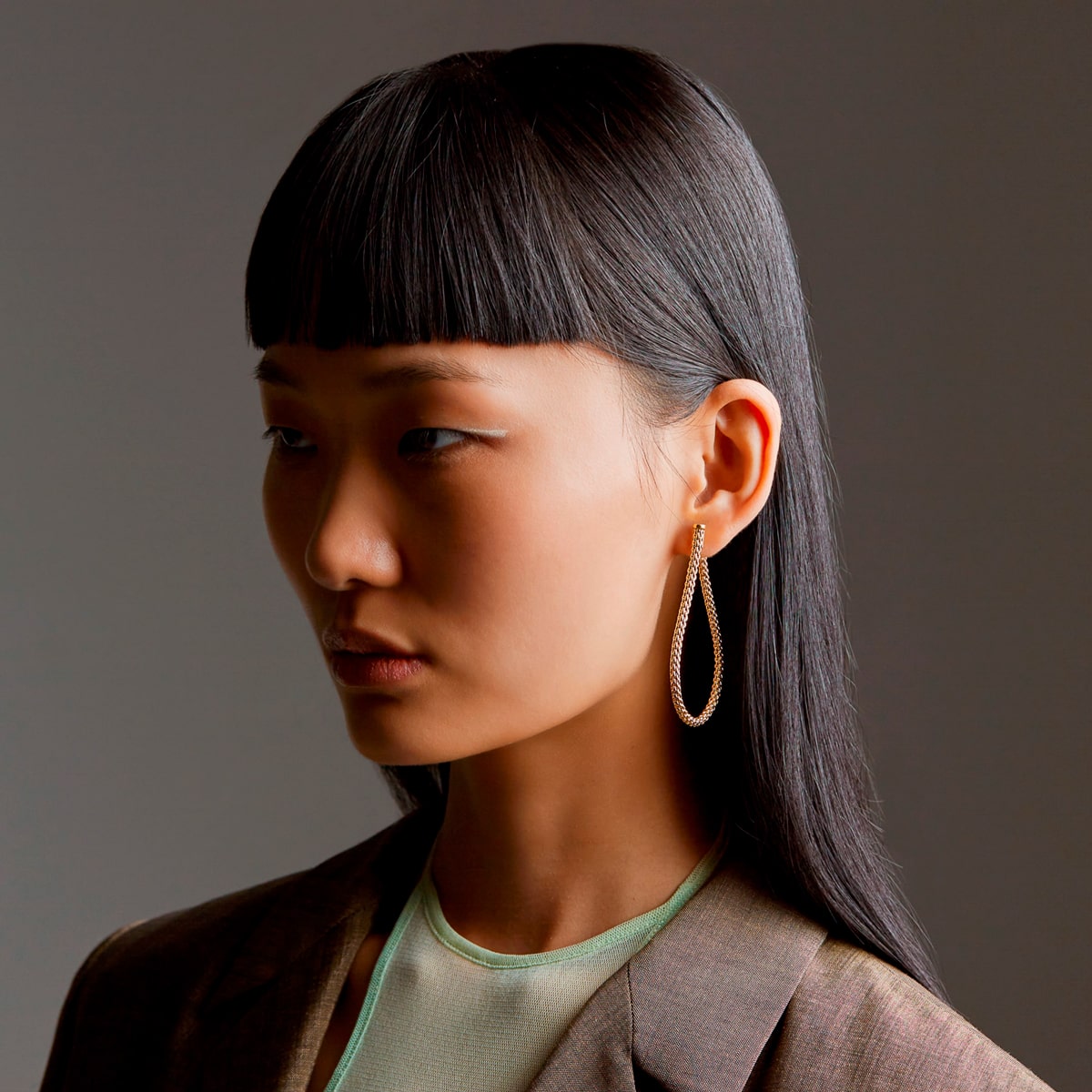 2025 © FOPE Eine Frau mit glattem schwarzem Haar und stumpfem Pony trägt auffällige FOPE-Ohrringe - große, tropfenförmige Reifen - und einen braunen Blazer über einem hellgrünen Oberteil, wobei sie vor einem schlichten neutralen Hintergrund nach unten und in die Ferne schaut.