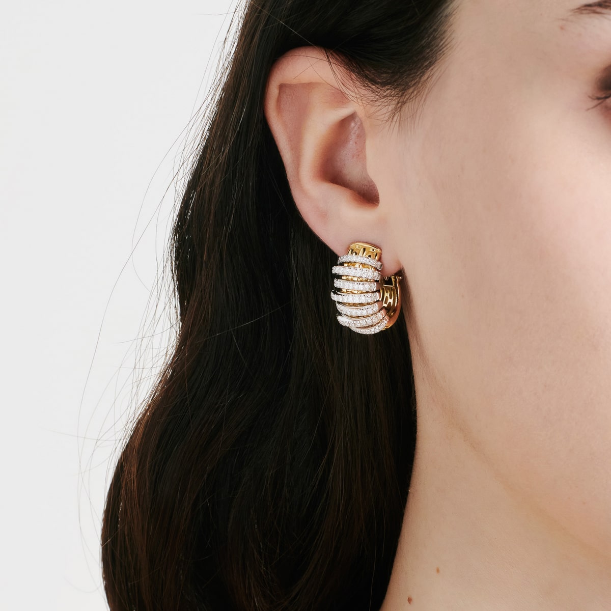 2025 © FOPE Nahaufnahme eines Frauenohrs, das FOPE Ohrringe trägt - goldene, mit Diamanten besetzte Ringe mit Rippenmuster. Ihr dunkles Haar ist hinter ihr Ohr gesteckt, vor einem schlichten, hellen Hintergrund.