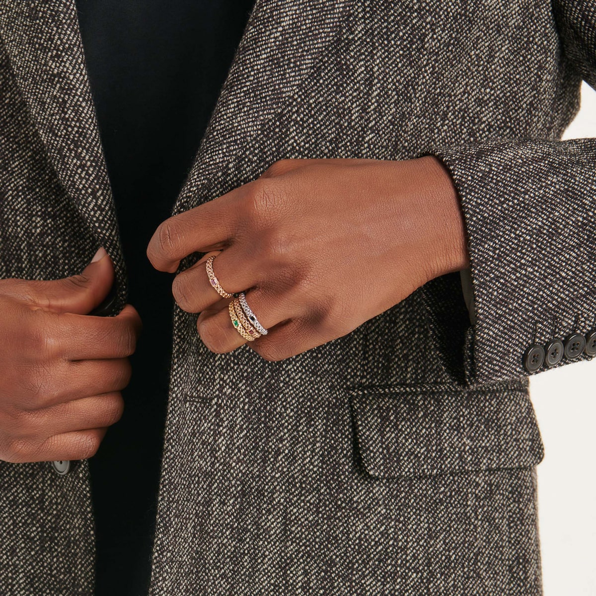 2025 © FOPE Eine Person, die einen strukturierten grauen Blazer und ein schwarzes Oberteil trägt, rückt das Revers zurecht. An ihrem Finger befinden sich drei FOPE-Ringe, die mit verschiedenen Metallen und Edelsteinen besetzt sind. Der Ärmel des Blazers ist mit vier schwarzen Knöpfen verziert.