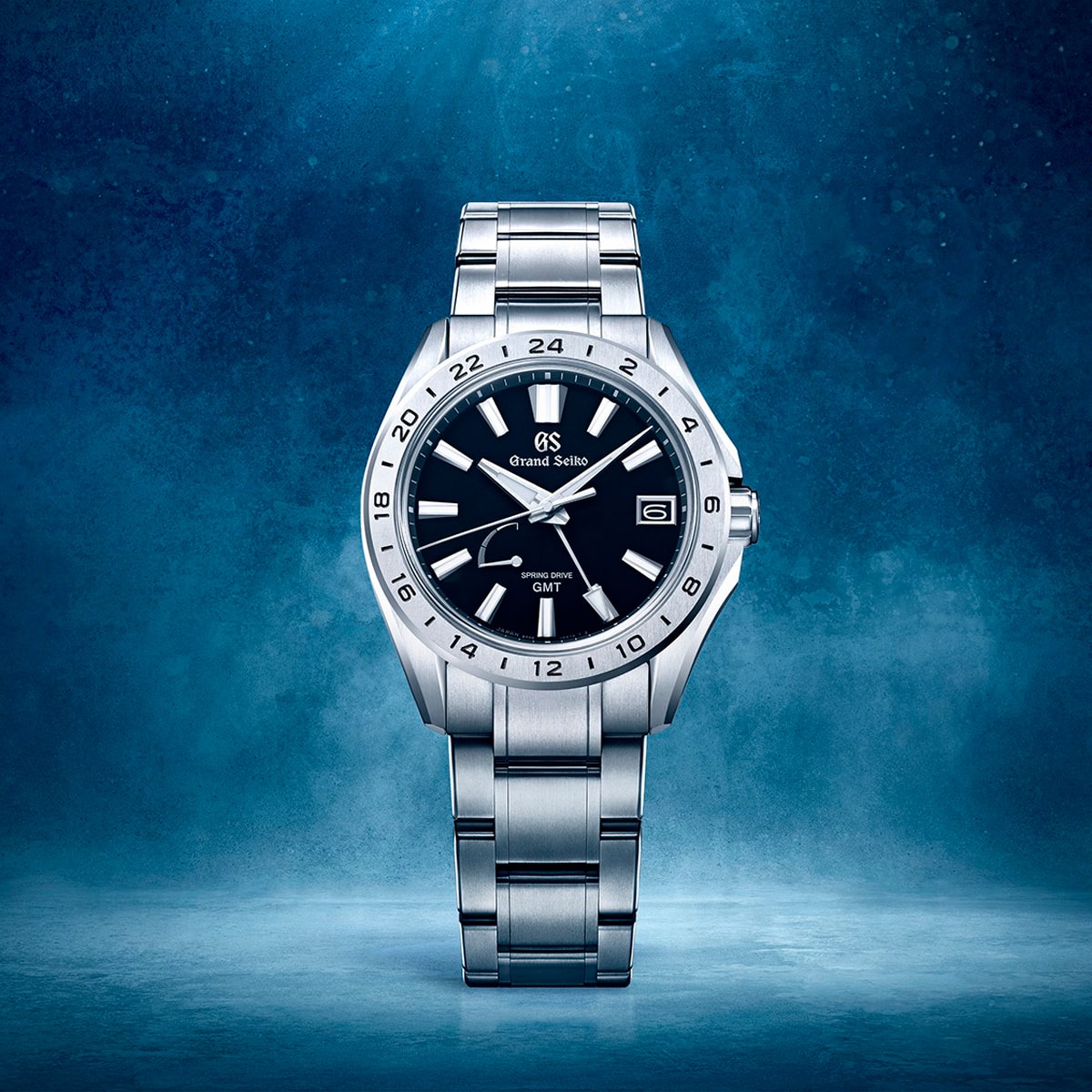2025 © GRAND SEIKO Eine Grand Seiko Evolution 9 Armbanduhr mit Edelstahlarmband und schwarzem Zifferblatt wird aufrecht vor einem strukturierten blauen Hintergrund präsentiert. Die Uhr verfügt über silberne Stundenmarkierungen, ein Datumsfenster und eine 24-Stunden-Lünette.