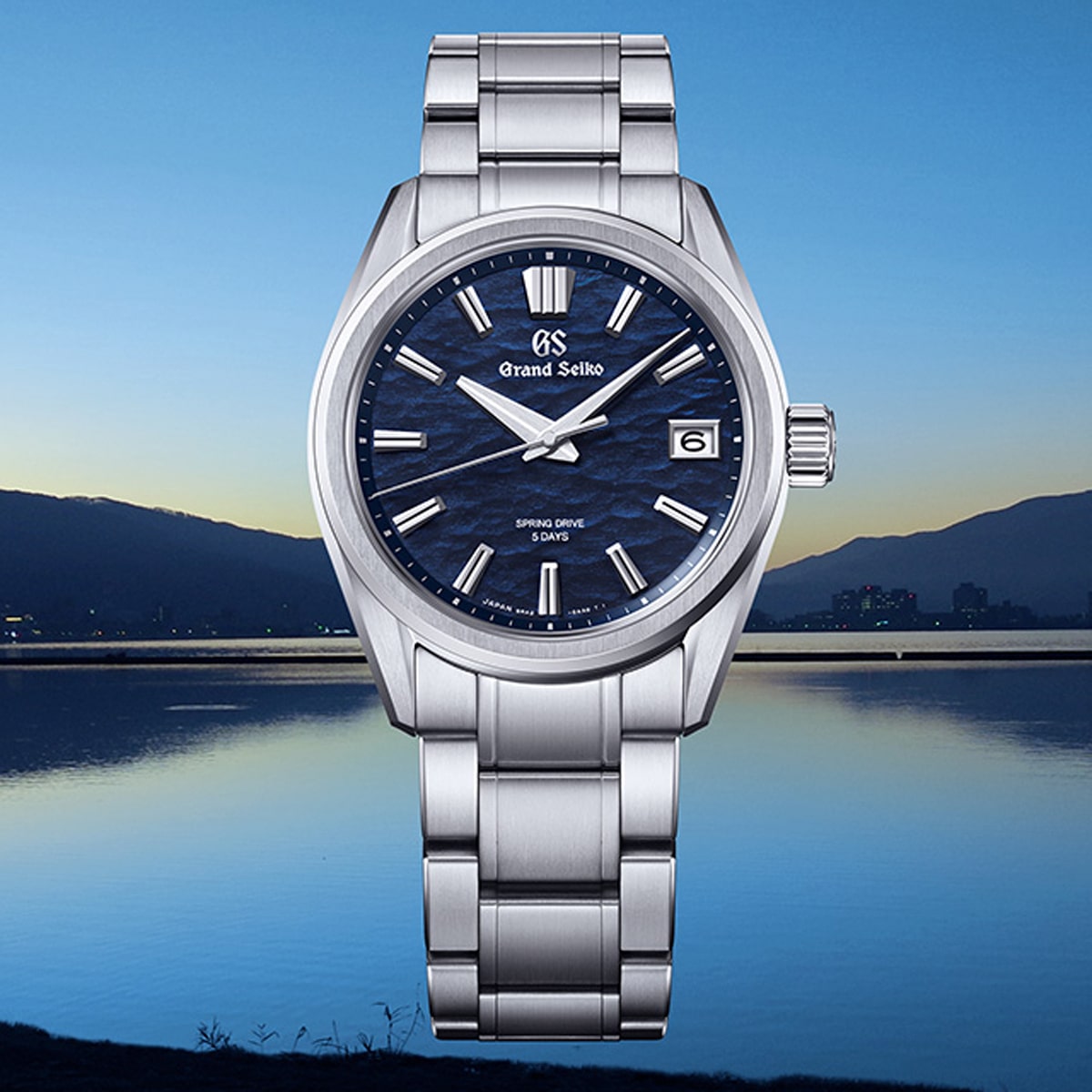 2025 © GRAND SEIKO Eine Grand Seiko Evolution 9 Edelstahlarmbanduhr mit strukturiertem blauem Zifferblatt und silbernen Indexen ist vor einer ruhigen See- und Berglandschaft in der Abenddämmerung zu sehen.