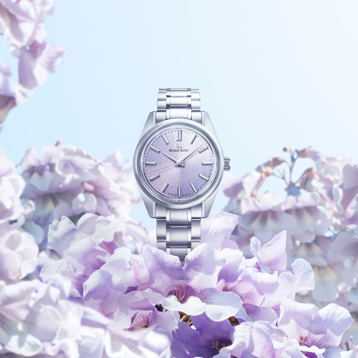 2025 © GRAND SEIKO Eine silberne Grand Seiko Heritage Armbanduhr mit hellviolettem Ziffernblatt steht inmitten von zarten, pastellvioletten Blumen unter einem hellblauen Himmel und macht einen frischen, eleganten und luxuriösen Eindruck.