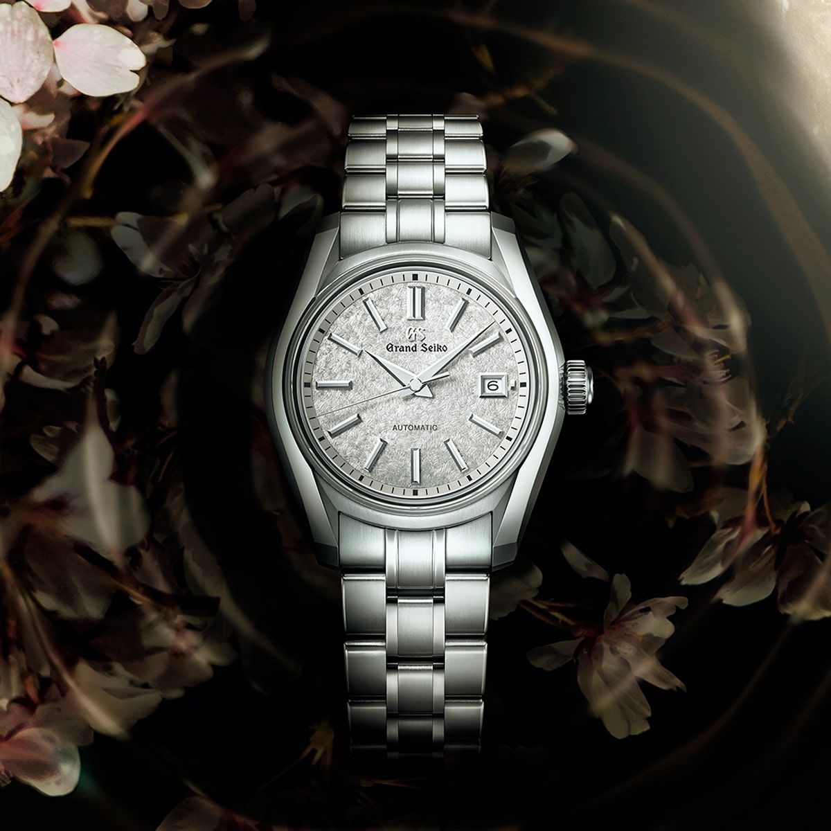 2025 © GRAND SEIKO Eine Grand Seiko Heritage Armbanduhr mit silbernem Metallarmband und einem strukturierten, hellgrauen Zifferblatt, das vor einem verschwommenen floralen Hintergrund mit sanften Rosa- und Brauntönen dargestellt ist.
