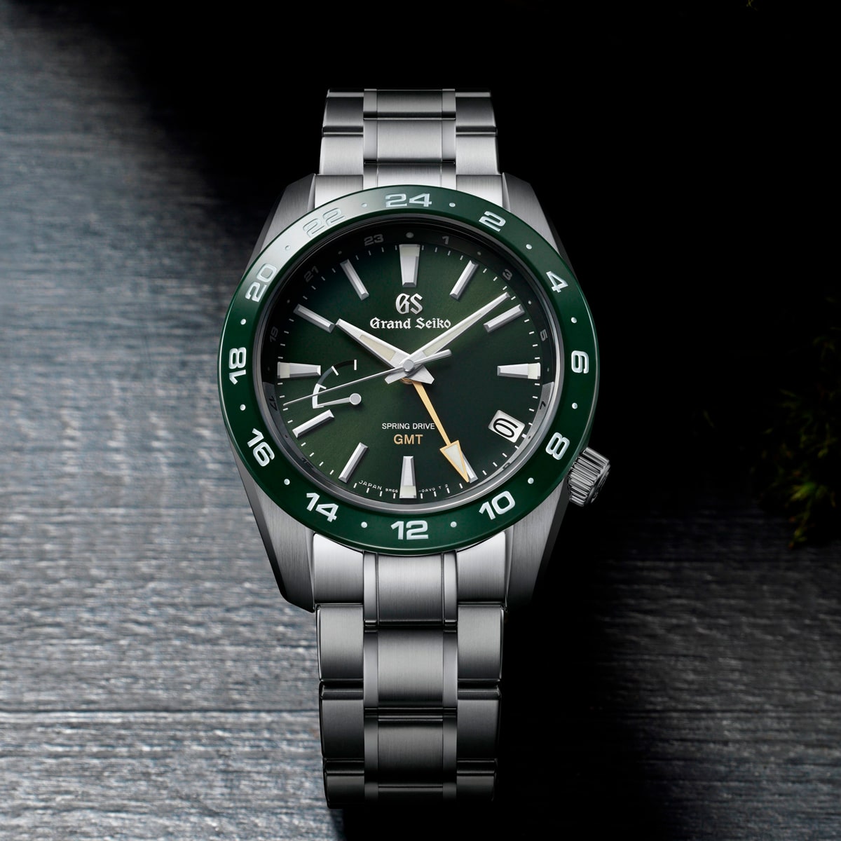 2025 © GRAND SEIKO Eine Grand Seiko Sport Armbanduhr mit silbernem Metallband, grüner Lünette und grünem Zifferblatt mit silbernen Stundenmarkierungen, Leuchtzeigern und einem GMT-Zeiger auf einer dunkel strukturierten Oberfläche.