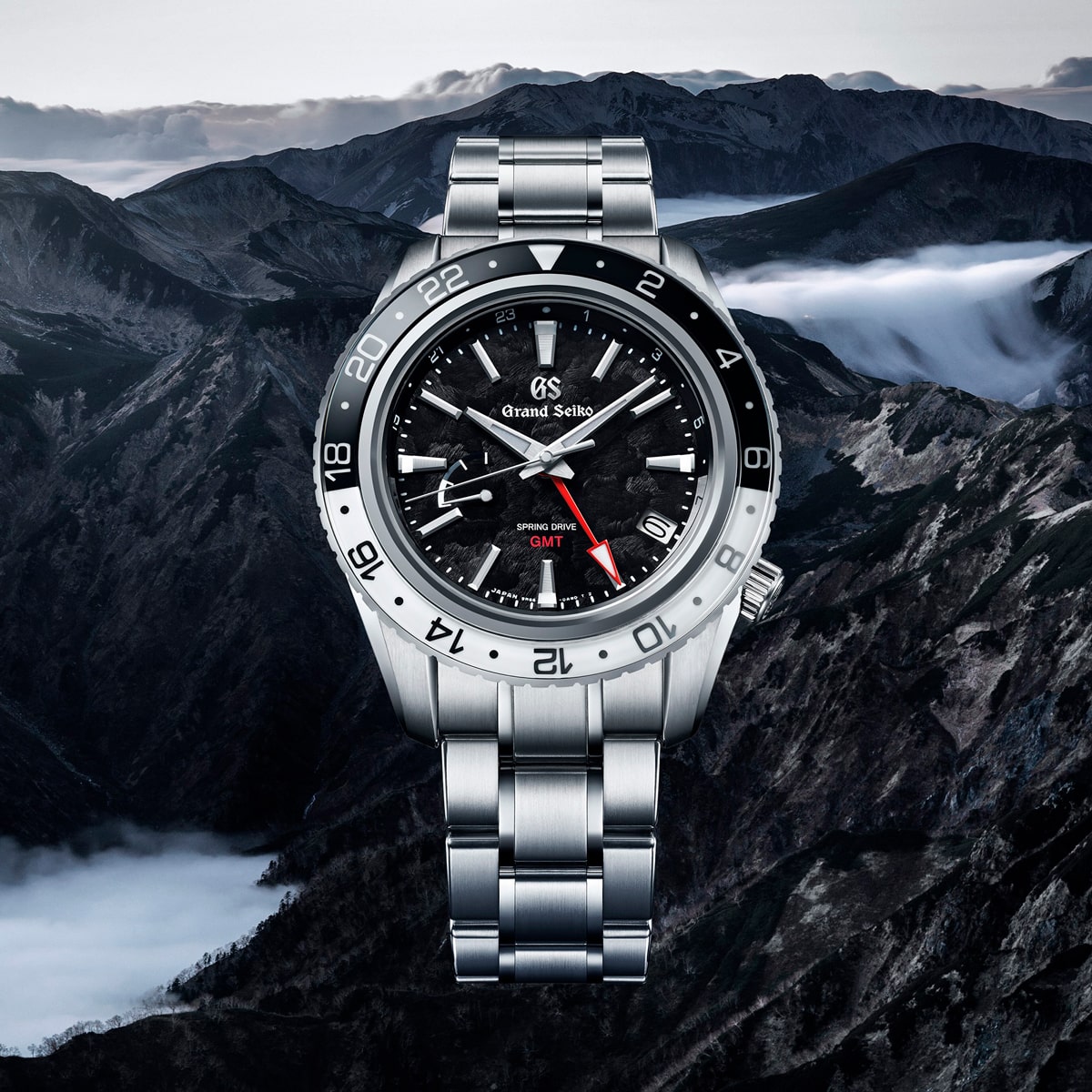 2025 © GRAND SEIKO Im Vordergrund steht eine Grand Seiko Sport GMT-Armbanduhr aus Edelstahl mit schwarzem, strukturiertem Zifferblatt vor einer dramatischen Berglandschaft mit Nebel und schroffen Gipfeln.