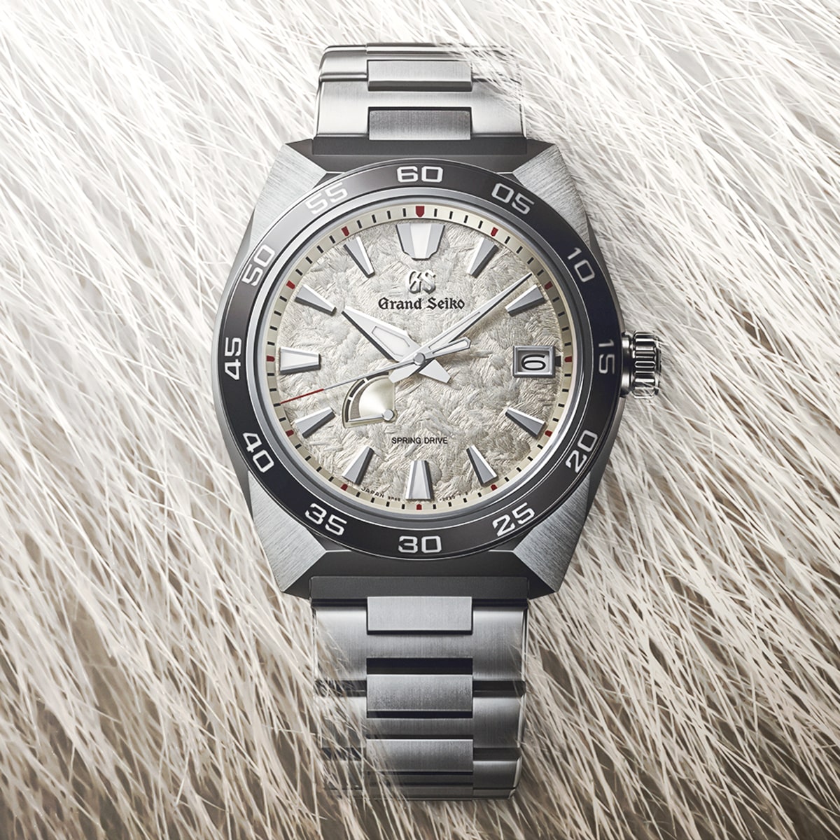 2025 © GRAND SEIKO Eine Grand Seiko Sport Armbanduhr mit silbernem Metallband und strukturiertem, hellem Zifferblatt ruht auf einer weichen, weißen, fellartigen Oberfläche. Die Uhr hat ein Datumsfenster und eine schwarze Lünette mit weißen Ziffern.