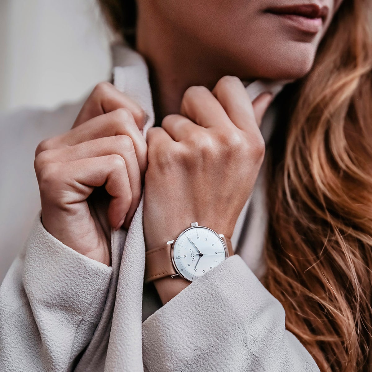 2025 © Junghans Nahaufnahme einer Person, die den Kragen eines hellbeigen Mantels festhält und eine minimalistische weiße Junghans Max Bill Armbanduhr mit einem hellbraunen Lederarmband trägt. Das lange, hellbraune Haar der Person ist auf einer Seite sichtbar.