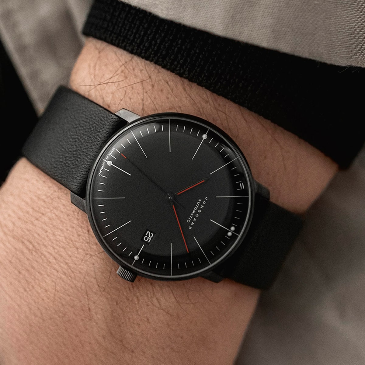 2025 © Junghans Nahaufnahme einer minimalistischen schwarzen Junghans Max Bill-Armbanduhr mit schwarzem Lederarmband am Handgelenk einer Person. Das Zifferblatt zeigt dünne weiße Stundenmarkierungen, zwei rote Zeiger und ein Datumsfenster bei 3 Uhr unter einer beigen Hülle.
