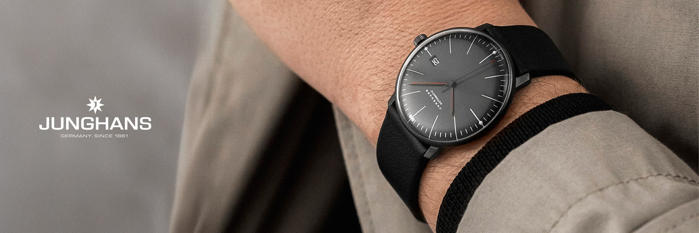 2025 © Junghans Nahaufnahme einer Person, die eine minimalistische Junghans Max Bill-Armbanduhr mit schwarzem Armband und schlichtem grauem Zifferblatt trägt. Das Junghans-Logo und "GERMANY. SINCE 1861" erscheinen auf der linken Seite des Bildes.