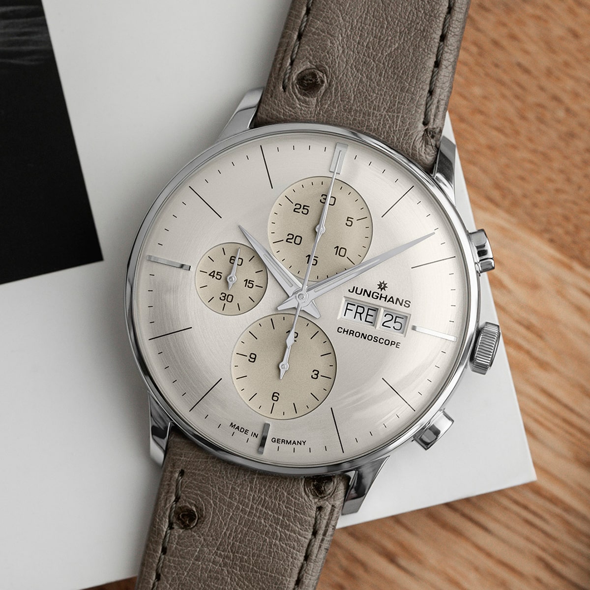 2025 © Junghans Eine silberne Junghans Meister Chronoscope Armbanduhr mit beigefarbenem Zifferblatt, drei Hilfszifferblättern, Tagesanzeige und braunem Lederarmband ruht auf einer Holzfläche neben einem weißen und schwarzen Buch.