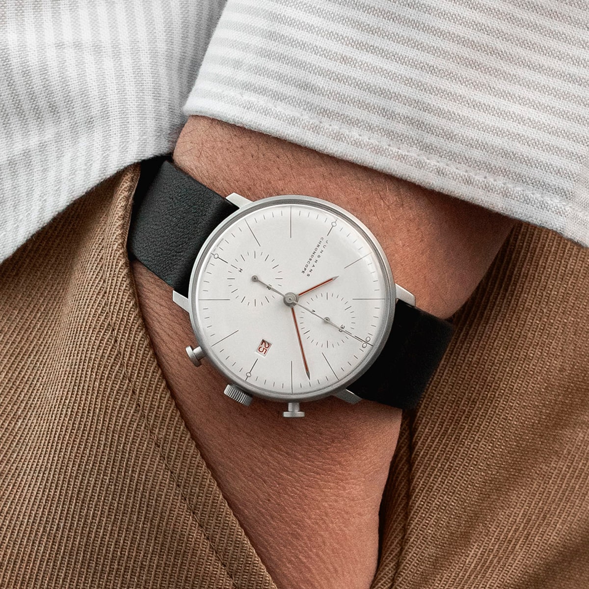2025 © Junghans Nahaufnahme einer minimalistischen Junghans Meister-Armbanduhr mit weißem Zifferblatt, zwei Hilfszifferblättern und schwarzem Lederarmband, getragen am Handgelenk einer Person, die ein gestreiftes Hemd und eine braune Hose trägt.
