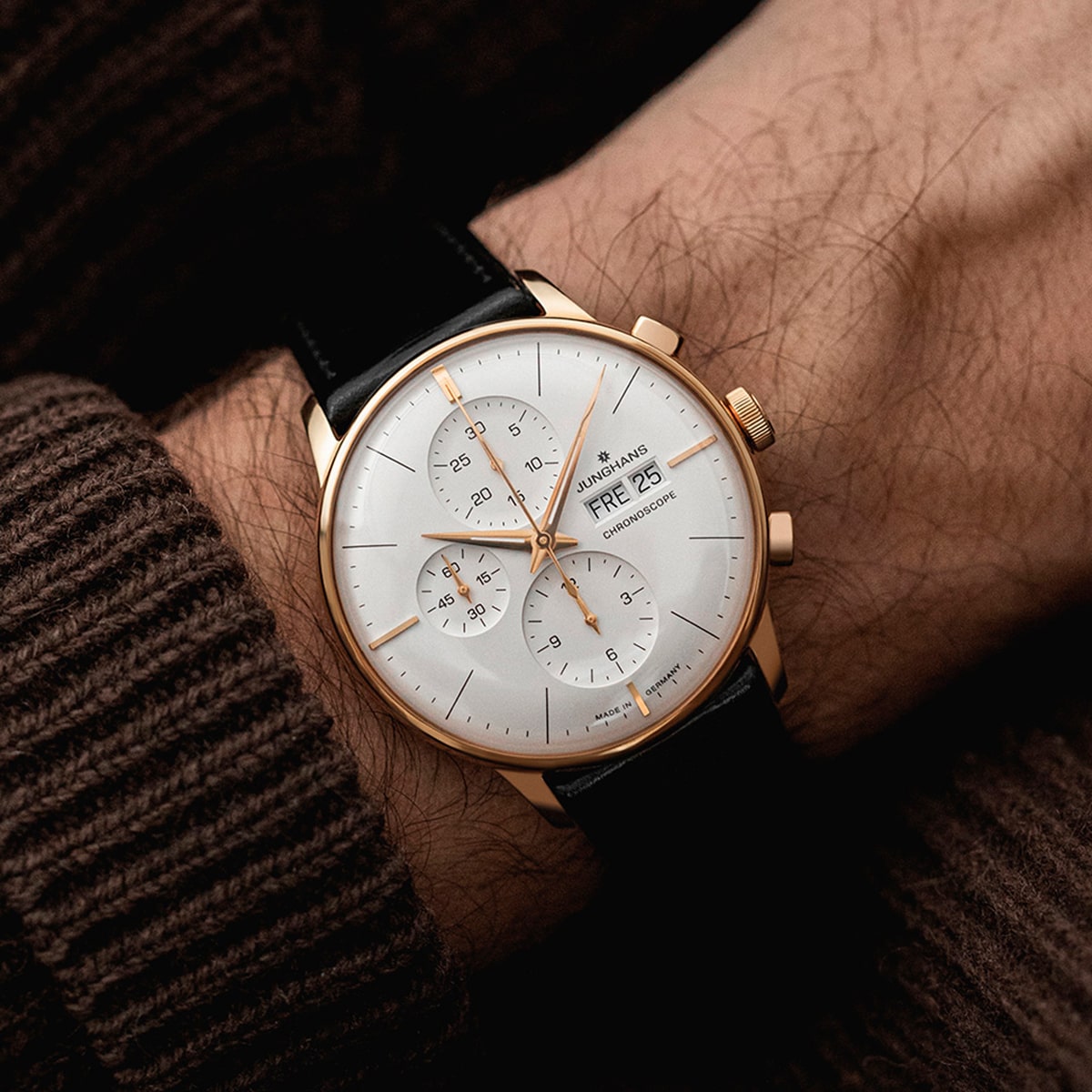 2025 © Junghans Eine Person, die einen braunen Pullover trägt, zeigt eine gold-weiße Junghans Meister-Armbanduhr mit schwarzem Lederarmband. Das Zifferblatt zeigt drei Ziffernblätter und das Datum "FRI 25".