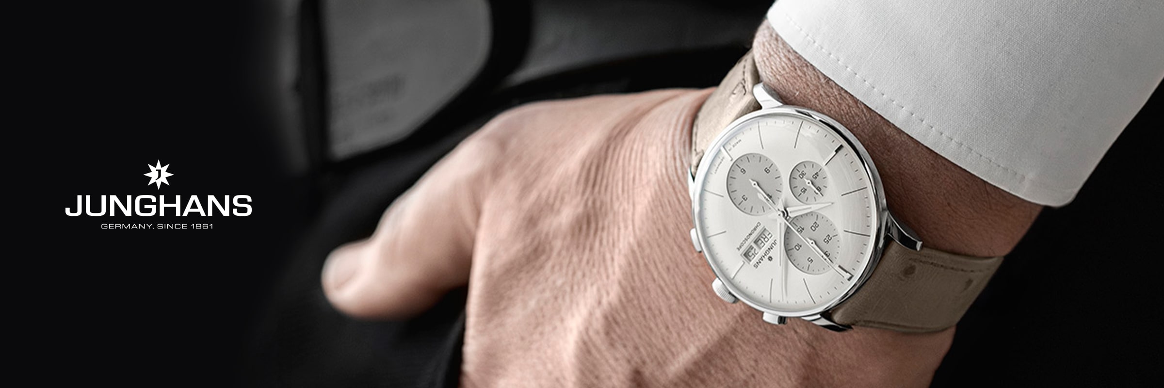 2025 © Junghans Nahaufnahme des Handgelenks einer Person, die eine Junghans Meister Chronographenuhr mit weißem Zifferblatt und beigem Lederarmband trägt. Die Person ist mit einem weißen Hemd bekleidet und hält einen schwarzen Gegenstand in der Hand; links erscheint das Junghans-Logo.