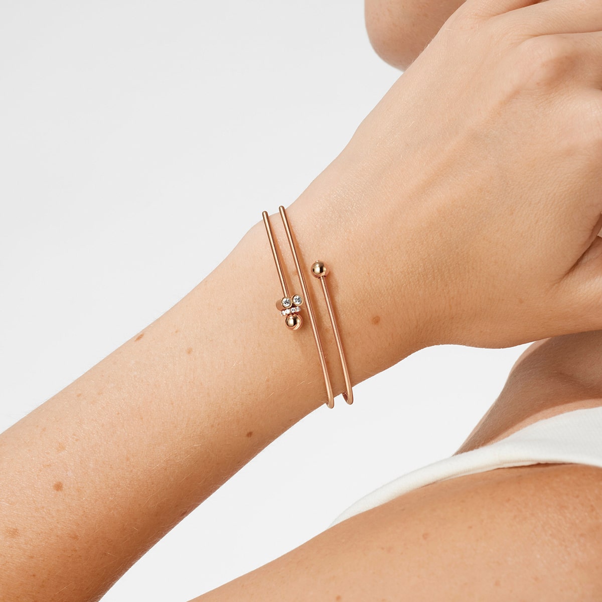 2025 © Niessing Nahaufnahme des Handgelenks einer Person mit zwei dünnen goldenen Armschmuck-Armbändern von Niessing - eines mit einer kleinen runden Perle, das andere mit drei zarten Silberanhängern - vor einem schlichten Hintergrund.