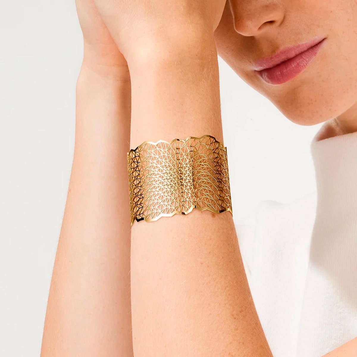 2025 © Niessing Eine Person, die ein breites, kunstvoll gemustertes goldenes Niessing-Armschmuck-Armband am Handgelenk trägt, wobei ein Teil ihres Gesichts und ein weißes Oberteil vor einem schlichten Hintergrund zu sehen sind.