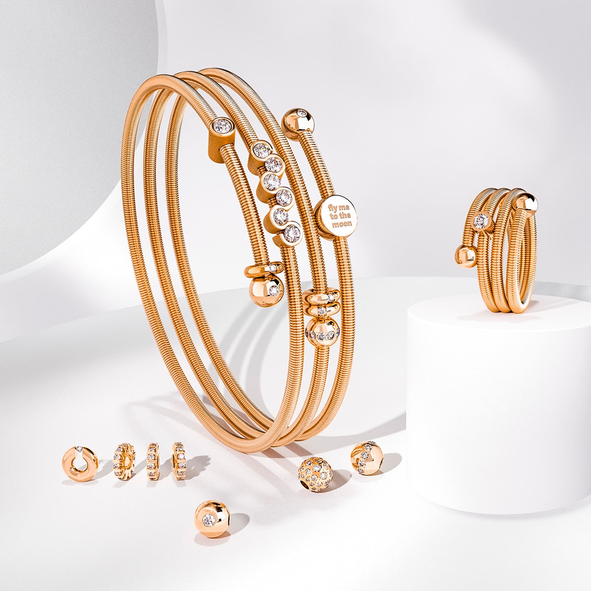 2025 © Niessing Ein Set aus Goldschmuck, bestehend aus Armreifen, Ringen und Ohrsteckern von Niessing Armschmuck mit eingebetteten klaren Edelsteinen, wird auf einer weißen Fläche mit weichen Schatten und einem minimalen Hintergrund präsentiert.