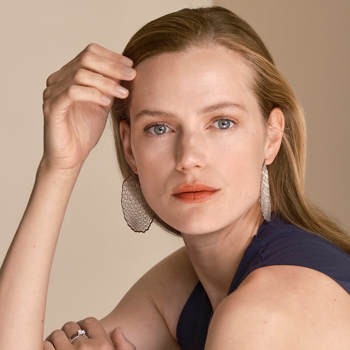2025 © Niessing Eine Frau mit heller Haut und blauen Augen blickt in die Kamera. Sie trägt große, strukturierte Niessing-Ohrschmuck-Ohrringe und ein ärmelloses marineblaues Oberteil. Sie hat hellbraunes Haar und berührt mit ihrer linken Hand leicht ihre Stirn.