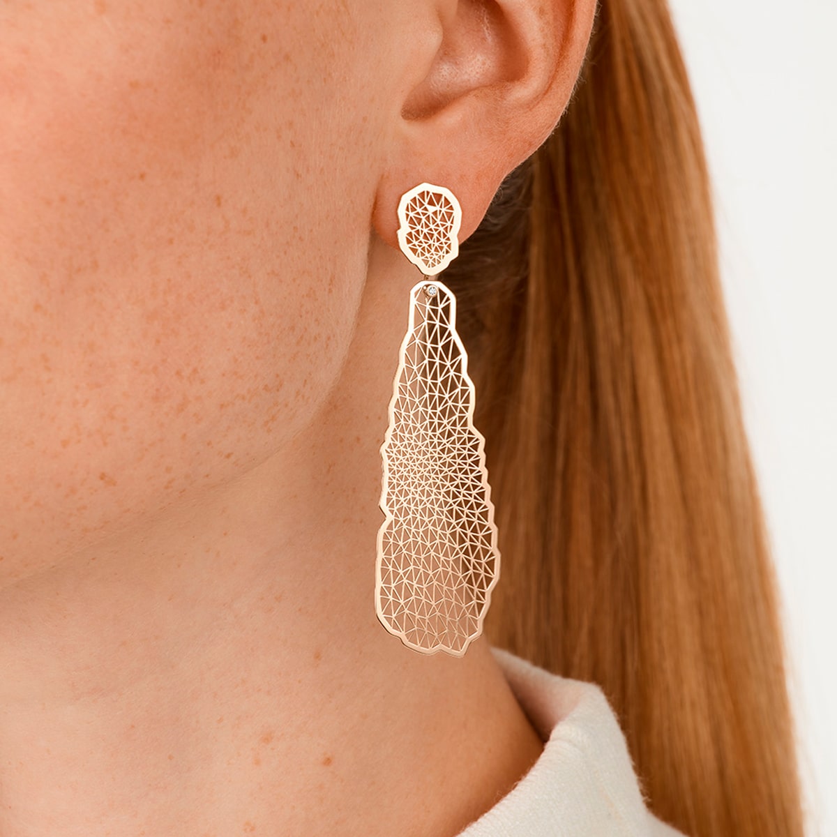 2025 © Niessing Nahaufnahme eines Frauenohrs mit einem großen, filigranen Niessing-Ohrschmuck aus Gold, der ein netzartiges geometrisches Muster aufweist. Ihr glattes Haar hat sie hinter das Ohr gesteckt, so dass ihre leicht sommersprossige Haut vor einem schlichten, hellen Hintergrund zum Vorschein kommt.