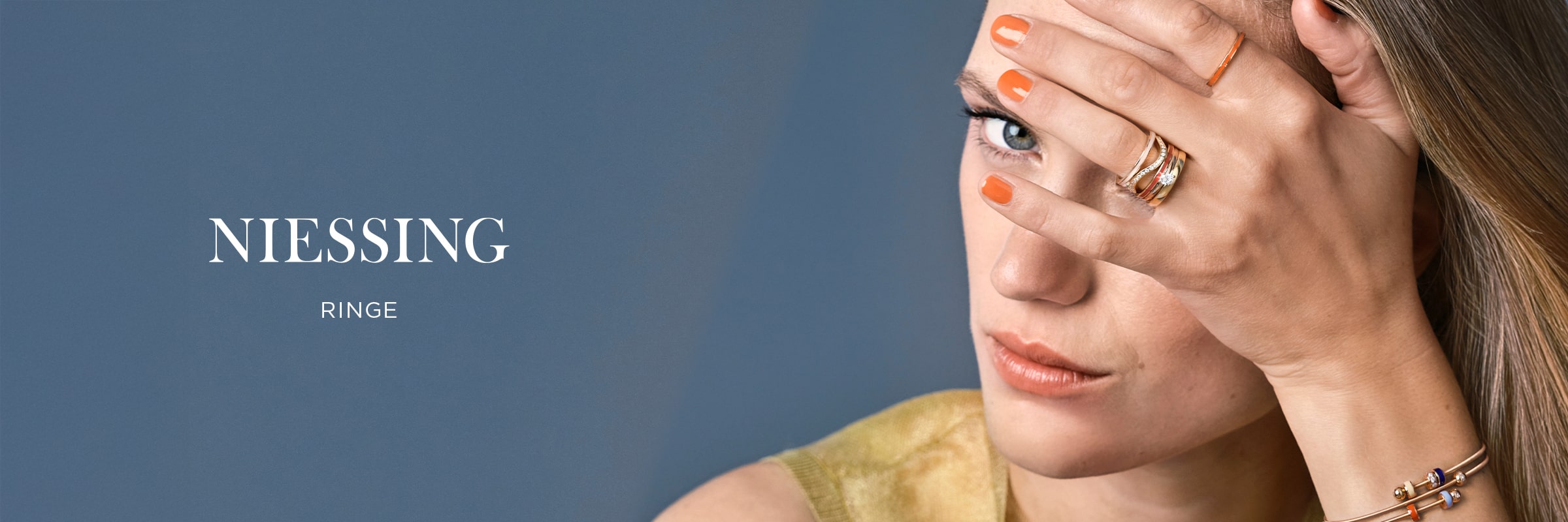 2025 © Niessing Eine Frau mit orangefarbenem Nagellack bedeckt ein Auge mit ihrer Hand, auf der mehrere goldene Niessing Ringe zu sehen sind. Der blaue Hintergrund hebt den Text "NIESSING RINGE" hervor.