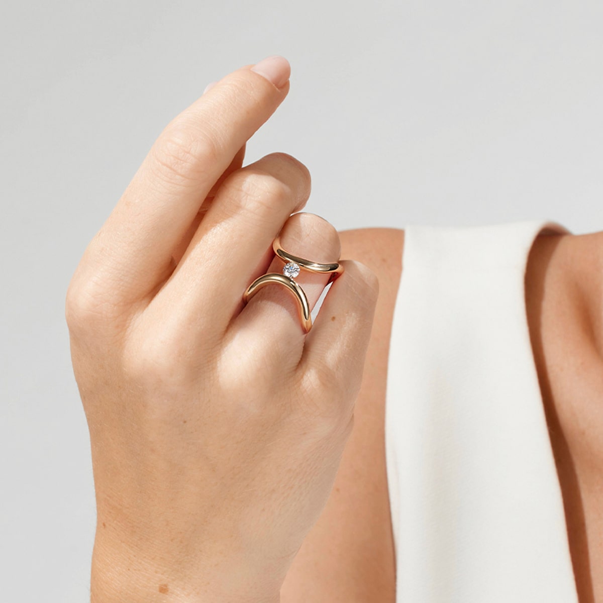 2025 © Niessing Eine Hand mit hellem Hautton trägt zwei goldene Ringe, darunter einen Niessing-Spannring mit einem kleinen Diamanten und ein schlichtes Band. Die Person ist mit einem weißen ärmellosen Oberteil vor einem neutralen Hintergrund bekleidet.