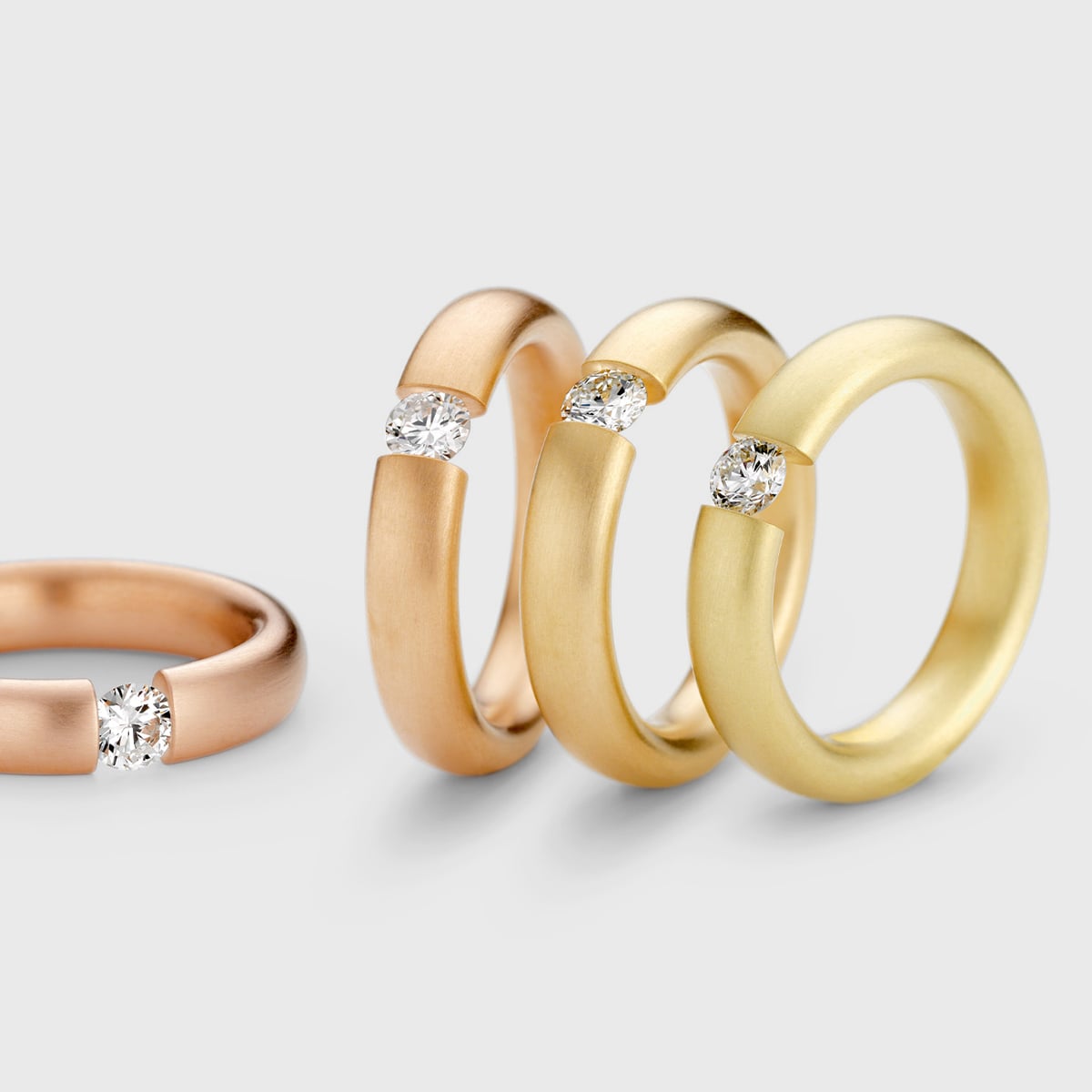 2025 © Niessing Vier Niessing Spannringe aus Gold und Roségold mit moderner, matter Oberfläche, jeder mit einem einzelnen runden Diamanten, der in einer Lücke an der Spitze gefasst ist. Die Ringe sind in einer Reihe auf einem hellgrauen Hintergrund angeordnet.