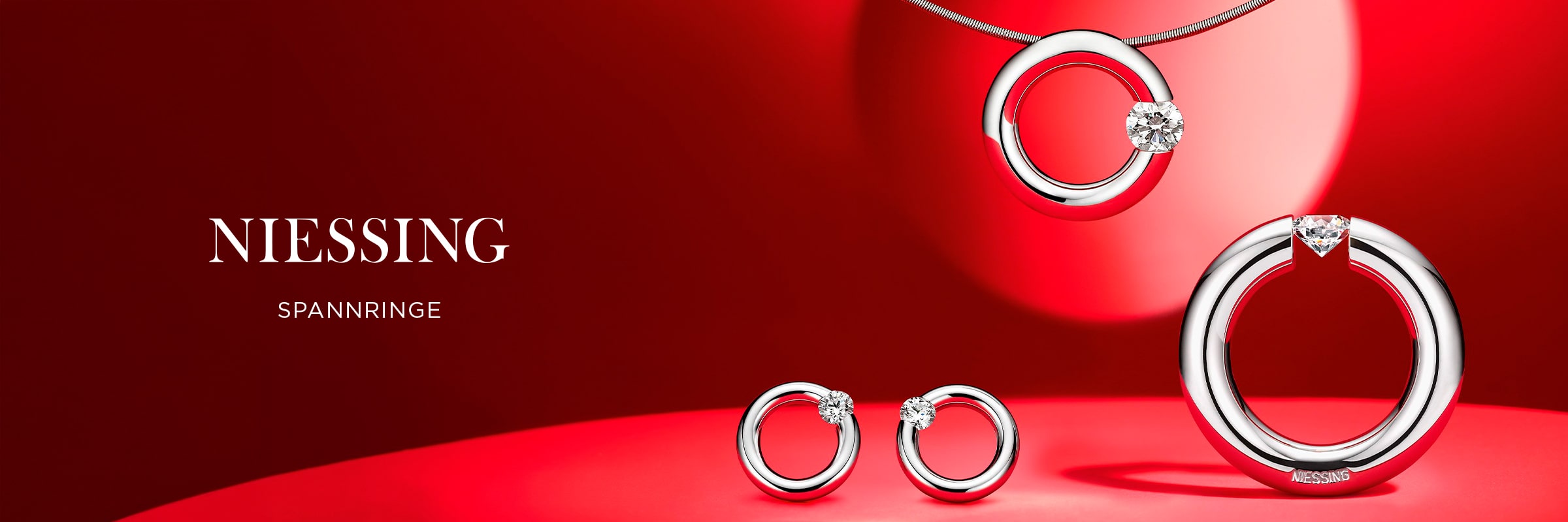 2025 © Niessing Kreisförmiger Silberschmuck mit einzelnen Diamanten - darunter eine Halskette, zwei Ohrringe und ein Ring - auf einem roten Hintergrund mit Scheinwerfern. Der Text lautet "NIESSING SPANNRINGE" und stellt die elegante Niessing Spannringe Kollektion vor.