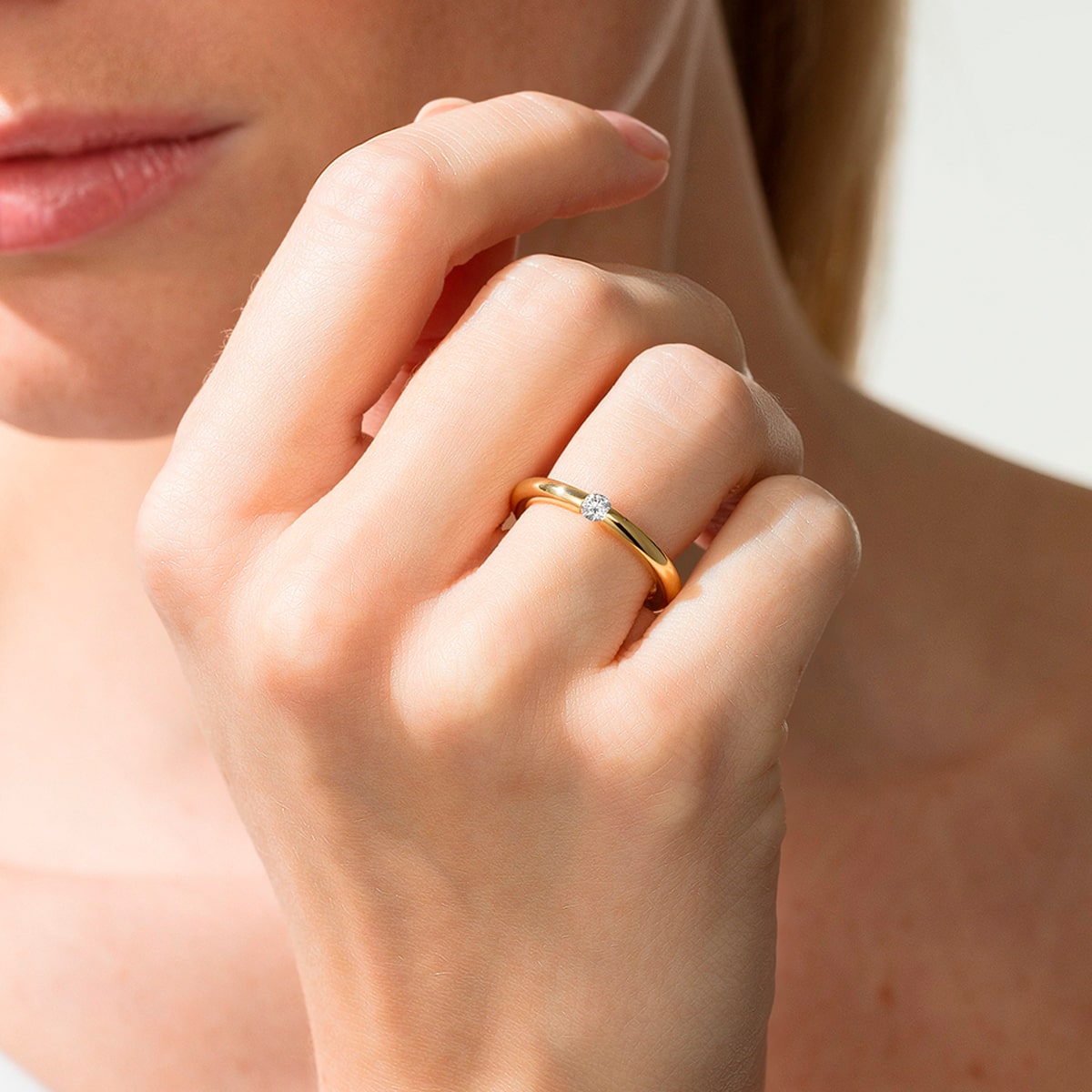 2025 © Niessing Eine Nahaufnahme der Hand einer Person, die einen goldenen Niessing Verlobungsring mit einem kleinen Diamanten am Ringfinger trägt. Die Lippen und das untere Gesicht der Person sind teilweise im Hintergrund zu sehen.