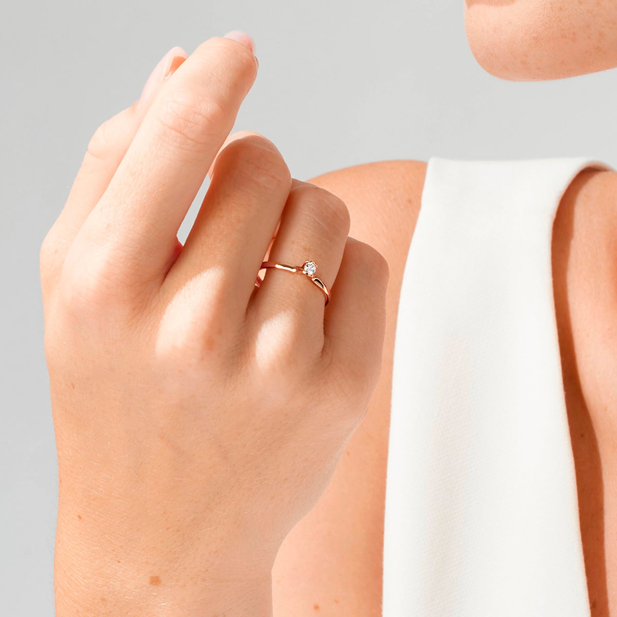2025 © Niessing Nahaufnahme einer Hand, die einen zarten Goldring mit einem kleinen Diamanten trägt, der an Niessing Verlobungsringe erinnert. Die Person ist mit einem ärmellosen weißen Oberteil vor einem hellen Hintergrund bekleidet.
