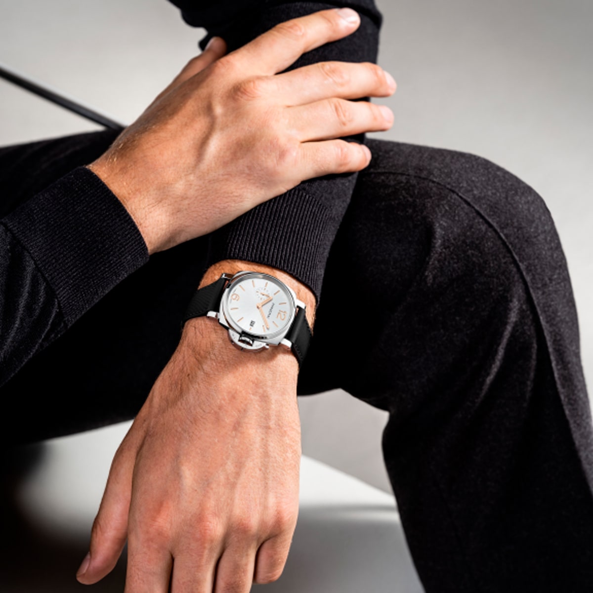 2025 © Panerai Eine Person, die ein schwarzes langärmeliges Hemd und eine Hose trägt, sitzt und zeigt eine Nahaufnahme ihres linken Handgelenks, das mit einer modernen silber-schwarzen Panerai Luminor Due Armbanduhr mit einem auffälligen weißen Zifferblatt geschmückt ist.