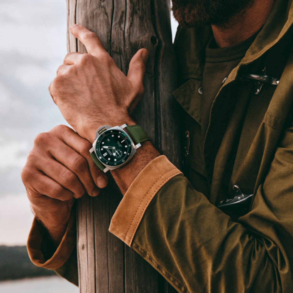2025 © Panerai Ein Mann in grüner Jacke lehnt mit verschränkten Armen an einem Holzpfosten. Er trägt eine Panerai Submersible mit grünem Armband; das Foto konzentriert sich auf seine Uhr und seine Zeiger. Der Hintergrund im Freien ist leicht unscharf.